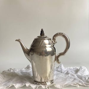 Può includere: Una caffettiera color argento con manico e beccuccio curvi. La caffettiera ha un coperchio decorativo con un pomello scuro. La caffettiera è su un panno bianco. La caffettiera è probabilmente vintage.