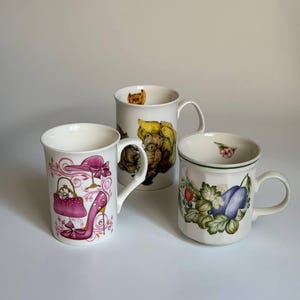May include: Three white ceramic mugs. The left mug features pink illustrations of a handbag, a high-heeled shoe, and a hat. The center mug displays a brown teddy bear illustration. The right mug shows a colorful fruit pattern with plums and berries.