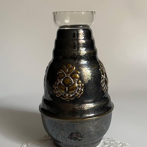 May include: A dark metallic vase with gold detailing and a clear glass top. The vase has embossed floral designs and a rounded shape. The base features a detailed pattern, and the overall style is vintage. The height is approximately 20 cm.