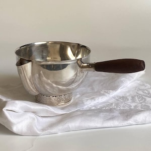 Può includere: Una piccola casseruola color argento con un manico in legno marrone scuro. La pentola ha un beccuccio e poggia su un tessuto bianco con un motivo floreale. La base della pentola presenta un design decorativo inciso.