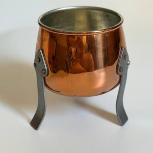 Op de afbeelding: Een kleine, ronde koperen pot met een gepolijste metalen afwerking en een zilverkleurige binnenkant. De pot wordt ondersteund door drie gebogen, matgrijze metalen poten die met kleine, zichtbare klinknagels aan de buitenkant zijn bevestigd.