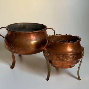 May include: Two copper vessels with metallic finishes. The larger bowl features two side handles and three curved legs. The smaller vessel has a scalloped rim, a decorative embossed band around the centre, and three slender metal legs.