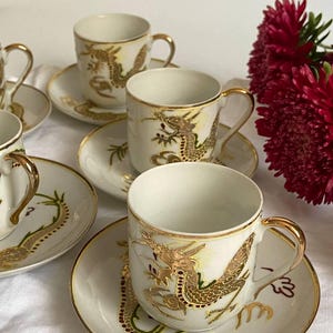 Può includere: Un set di tazze e piattini in porcellana bianca con dettagli dorati a forma di drago. Le tazze hanno manici dorati e sono esposte su una superficie bianca. I piattini presentano un design a drago coordinato.