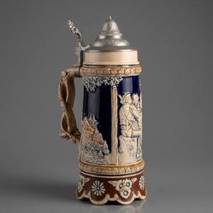 Peut inclure: Une chope à bière en céramique avec un couvercle métallique à charnière. La chope présente un corps bleu foncé avec des sculptures en relief de personnages et d'un paysage. La poignée est de couleur marron clair et la base est ornée de motifs floraux.