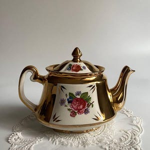 Puede incluir: Una tetera vintage con acabado dorado y un diseño floral con rosas rojas y flores azules. La tetera está sobre un tapete de encaje blanco. El asa y el pico también son dorados.