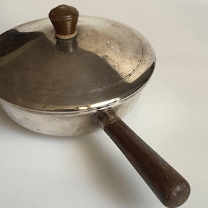 May include: A small, silver-colored saucepan with a lid and a long, dark brown wooden handle. The lid has a matching wooden knob. The pan is round and appears to be vintage or antique.