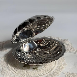 May include: A silver-toned metal trinket box shaped like an open seashell. The exterior and interior surfaces feature a polished, reflective finish with moulded ridges and a scalloped edge. The two halves are connected by a small metal hinge.