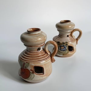 May include: Two ceramic oil lamps with a beige and brown glazed finish. Each lamp features a rounded base with horizontal ridges, a side handle, a small square opening, and a painted design of either a red apple or a blue flower.