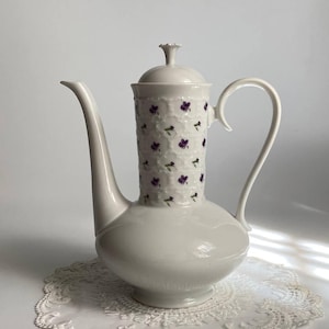Puede incluir: Una cafetera de porcelana blanca con un pico largo y un asa curvada. La parte superior cilíndrica está decorada con un patrón de flores moradas. La cafetera está sobre un tapete de encaje blanco.