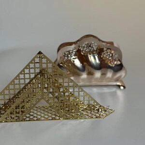 May include: A gold-toned metal napkin holder with a triangular shape and a lattice cutout pattern, positioned in front of a silver-toned metal toast rack featuring a scalloped top edge and decorative floral cutouts along the sides.