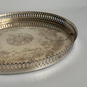 Könnte beinhalten: Ein rundes silberfarbenes Metalltablett mit einem komplexen gravierten Blumenmuster auf der Basis und einem erhöhten Rand mit einem sich wiederholenden durchbrochenen Ausschnittdesign. Die Oberfläche hat ein poliertes Finish.