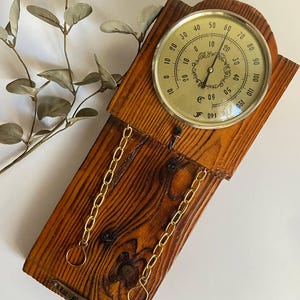May include: A vintage wooden thermometer with a brass-colored face and a temperature gauge. The thermometer is mounted on a stained wooden plank with decorative gold chains. The text "Ru-Mallorca" is visible.