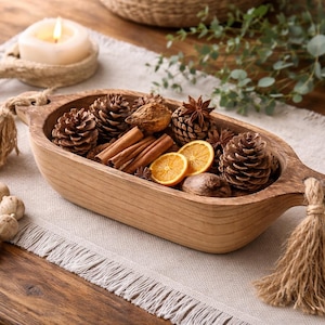May include: A wooden decorative bowl filled with pine cones, cinnamon sticks, orange slices, and star anise. A lit candle sits on a small, round, woven coaster. A beaded garland and a tassel are also present.