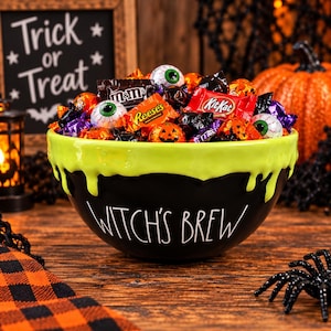 May include: A black ceramic bowl with the text Witch's Brew printed in white, featuring a lime green drip design around the rim. The bowl is filled with assorted Halloween candies, including chocolate bars, orange pumpkin-shaped candies, and eyeball-shaped treats.