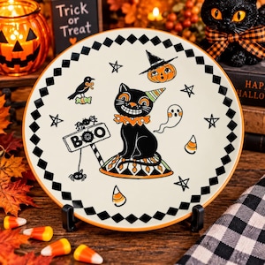 May include: A round ceramic plate with a cream background and a black and white diamond border. The center features a black cat wearing a party hat and ruffled collar, surrounded by a ghost, a pumpkin, a crow, stars, and a sign that reads BOO.