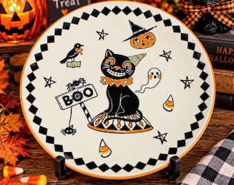 May include: A round ceramic plate with a cream background and a black and white diamond border. The center features a black cat wearing a party hat and ruffled collar, surrounded by a ghost, a pumpkin, a crow, stars, and a sign that reads BOO.