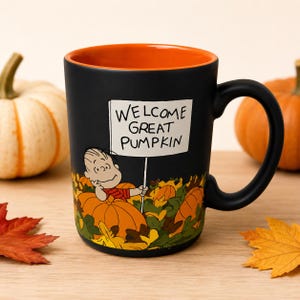 May include: A matte black ceramic mug with a bright orange interior and a handle. The exterior features a graphic of a cartoon character holding a sign that reads "WELCOME GREAT PUMPKIN" amidst a patch of orange pumpkins and green foliage.