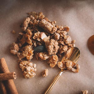 May include: A close-up shot of homemade granola with almonds, pumpkin seeds, and other ingredients. A gold spoon scoops up a portion of the granola. Cinnamon sticks are on the left side of the image.