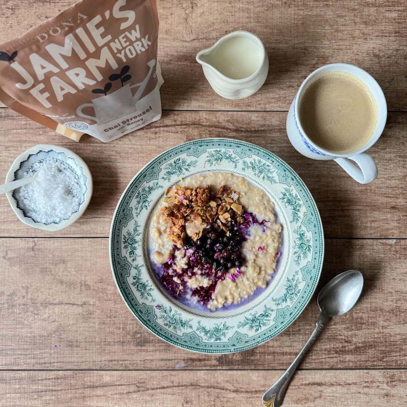 May include: A breakfast scene featuring a bowl of oatmeal topped with berries and granola, a cup of coffee, a small creamer, and a dish of salt. A bag of "Jamie's Farm New York" Chai Streusel is also present. The items are arranged on a wooden surface.