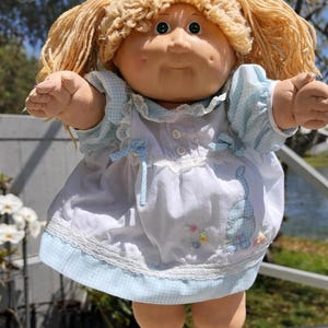 Puede incluir: Muñeca vintage Cabbage Patch Kids con cabello rubio de hilo y una cara redonda con mejillas rosadas. La muñeca viste un vestido blanco con ribete de cuadros azules y una aplicación de gato azul. Los brazos y las piernas de la muñeca están extendidos.