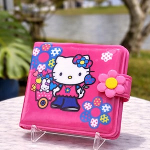 May include: A pink Hello Kitty notebook with a flower clasp. The cover features Hello Kitty, flowers, and a small wagon. The notebook is displayed on a clear stand.