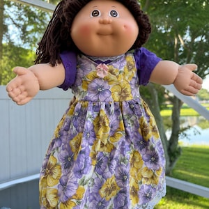 Könnte beinhalten: Eine Puppe mit dunkelbraunem Wollhaar, rundem Gesicht und rosigen Wangen. Sie trägt ein violettes Kurzarmshirt unter einem Kleid mit gelb-violettem Stiefmütterchen-Blumenmuster, verziert mit einer kleinen rosa Schleife und weißer Spitzenborte am Saum.