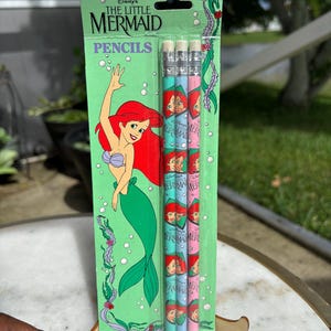 Op de afbeelding: Een verpakking van Disney's De Kleine Zeemeermin potloden. De potloden zijn lichtblauw en roze met Ariel's afbeelding. De verpakking is groen met Ariel's illustratie en de woorden "De Kleine Zeemeermin" en "Potloden".