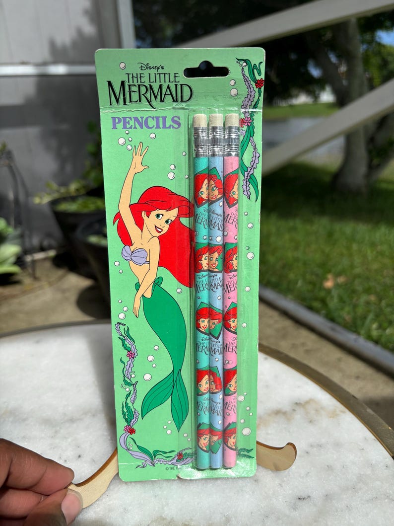 Op de afbeelding: Een verpakking van Disney's De Kleine Zeemeermin potloden. De potloden zijn lichtblauw en roze met Ariel's afbeelding. De verpakking is groen met Ariel's illustratie en de woorden "De Kleine Zeemeermin" en "Potloden".