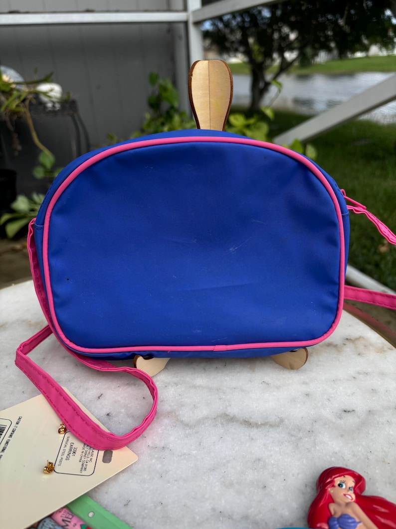 Op de afbeelding: Een blauwe handtas met roze rand en riem, met een houten schildpadontwerp. De tas heeft een afgeronde vorm en een roze riem. Een klein Ariel-beeldje is rechtsonder zichtbaar.
