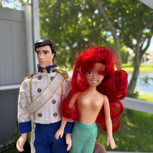 May include: Two dolls: a male doll in a white jacket with gold trim and blue pants, and a mermaid doll with red hair and a green tail. The dolls are held up in front of a blurred outdoor background.