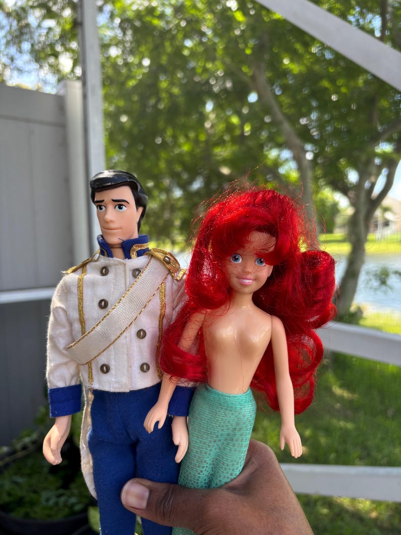 May include: Two dolls: a male doll in a white jacket with gold trim and blue pants, and a mermaid doll with red hair and a green tail. The dolls are held up in front of a blurred outdoor background.