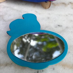 Op de afbeelding: Een turquoise compacte spiegel met een afgeronde ovale spiegel en een silhouetontwerp. De spiegel heeft een turquoise frame en een reflecterend oppervlak. De tekst "&copy; DISNEY" is zichtbaar aan de bovenkant van de spiegel.