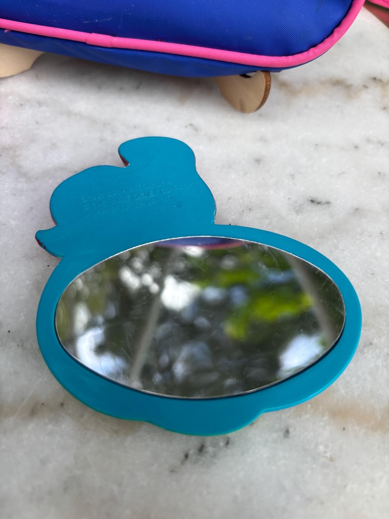 Op de afbeelding: Een turquoise compacte spiegel met een afgeronde ovale spiegel en een silhouetontwerp. De spiegel heeft een turquoise frame en een reflecterend oppervlak. De tekst "&copy; DISNEY" is zichtbaar aan de bovenkant van de spiegel.