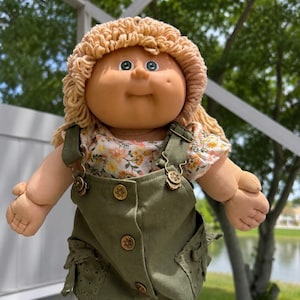 May include: A doll with light skin, dimples, and yarn hair in a light tan color. The doll wears a floral print top under olive green overalls featuring metal buttons and eyelet lace pockets. The doll has green eyes and a soft, rounded facial structure.