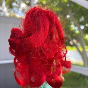 May include: Bright red doll hair, styled in voluminous curls. The hair is a vibrant shade of red, with a slightly tousled appearance. The hair is attached to a light green fabric base.