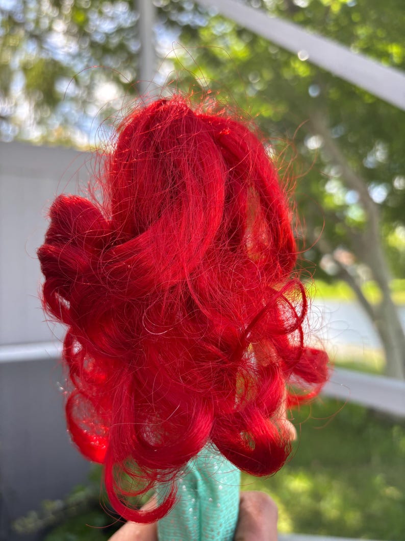 May include: Bright red doll hair, styled in voluminous curls. The hair is a vibrant shade of red, with a slightly tousled appearance. The hair is attached to a light green fabric base.