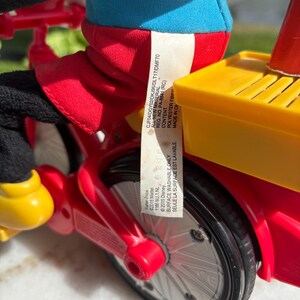 May include: A close-up of a red and yellow toy bicycle with black wheels. The toy has a blue and red figure riding it. The toy has a yellow seat and a red frame. A tag with text is attached to the toy.