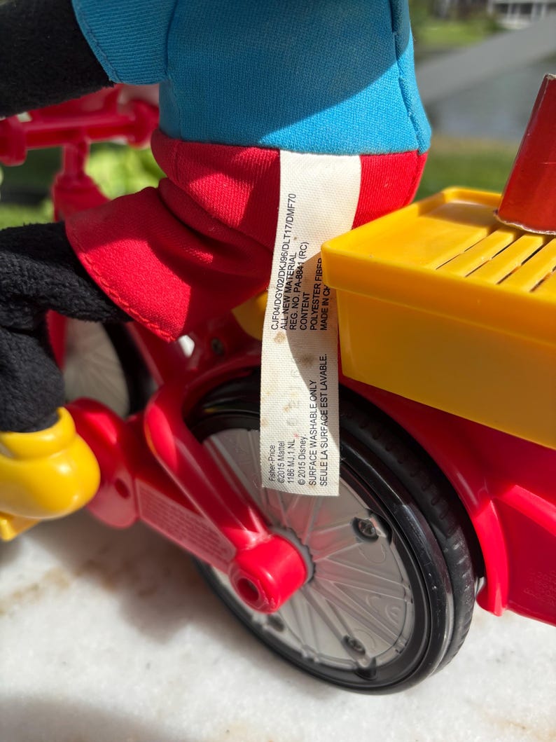 May include: A close-up of a red and yellow toy bicycle with black wheels. The toy has a blue and red figure riding it. The toy has a yellow seat and a red frame. A tag with text is attached to the toy.