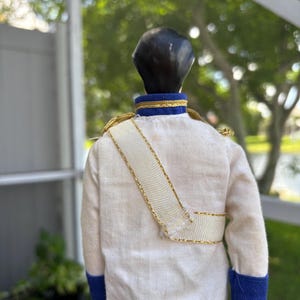 May include: A vintage doll wearing a white jacket with gold trim and a white sash. The jacket has blue cuffs and a blue and gold collar. The doll has black hair.