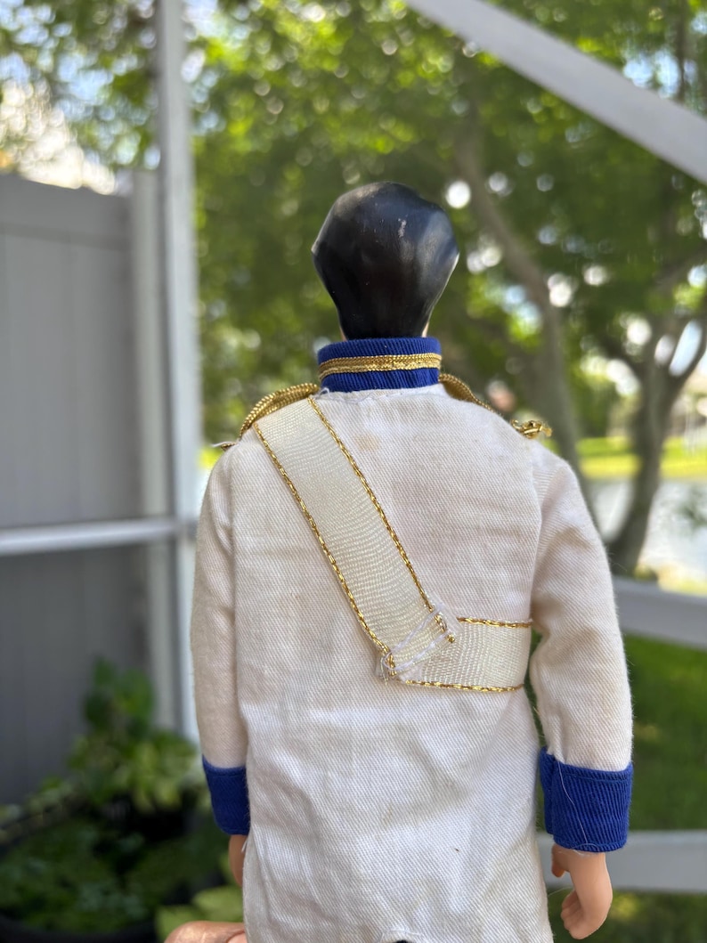 May include: A vintage doll wearing a white jacket with gold trim and a white sash. The jacket has blue cuffs and a blue and gold collar. The doll has black hair.
