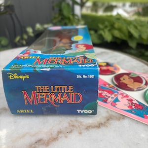 Pode incluir: Uma caixa retangular azul para uma boneca Ariel de A Pequena Sereia da Disney da Tyco, n&uacute;mero de estoque 1801, com ilustra&ccedil;&otilde;es de personagens. Ao lado da caixa h&aacute; broches circulares e uma folha de adesivos com imagens de Ariel e Sebasti&atilde;o.