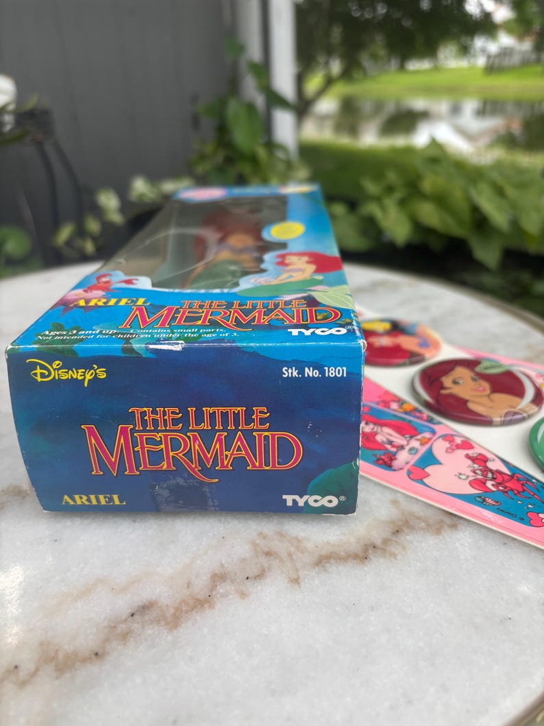 Pode incluir: Uma caixa retangular azul para uma boneca Ariel de A Pequena Sereia da Disney da Tyco, n&uacute;mero de estoque 1801, com ilustra&ccedil;&otilde;es de personagens. Ao lado da caixa h&aacute; broches circulares e uma folha de adesivos com imagens de Ariel e Sebasti&atilde;o.
