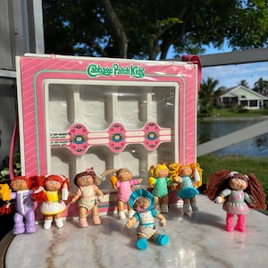 Puede incluir: Una colección de siete muñecas de plástico Cabbage Patch Kids con diversos atuendos y colores de cabello, exhibidas frente a un estuche rectangular rosa y blanco con el logotipo de Cabbage Patch Kids y etiquetas de nombre impresas.