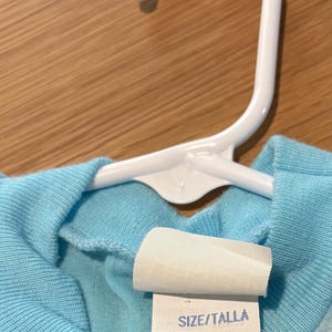 May include: A light blue children's shirt hanging on a white plastic hanger. The shirt has a tag with the size S (6/6X) and the text "MADE IN U.S.A."