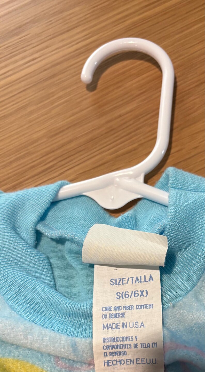 May include: A light blue children's shirt hanging on a white plastic hanger. The shirt has a tag with the size S (6/6X) and the text "MADE IN U.S.A."