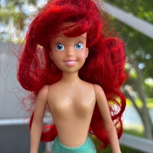 May include: A doll of Ariel, the Little Mermaid, with bright red hair and blue eyes. She wears a teal green mermaid tail. The doll has a smiling expression and is made of plastic.
