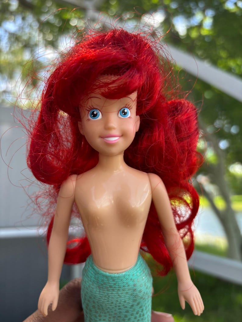 May include: A doll of Ariel, the Little Mermaid, with bright red hair and blue eyes. She wears a teal green mermaid tail. The doll has a smiling expression and is made of plastic.