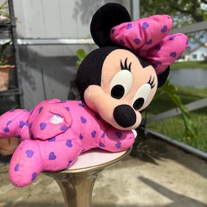 Pu&ograve; includere: Una bambola di peluche di Minnie Mouse in posizione sdraiata. La bambola &egrave; principalmente rosa con motivi a cuore viola e un grande fiocco. La bambola ha orecchie nere, un naso nero e grandi occhi bianchi. La bambola &egrave; appoggiata su un piedistallo dorato.