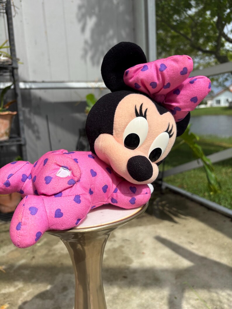 Pu&ograve; includere: Una bambola di peluche di Minnie Mouse in posizione sdraiata. La bambola &egrave; principalmente rosa con motivi a cuore viola e un grande fiocco. La bambola ha orecchie nere, un naso nero e grandi occhi bianchi. La bambola &egrave; appoggiata su un piedistallo dorato.