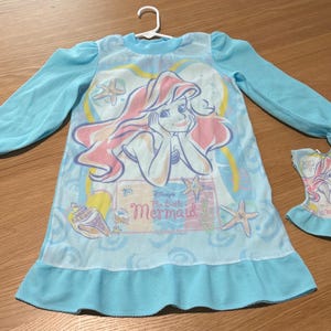 May include: A light blue nightgown featuring a graphic of Ariel from Disney's The Little Mermaid. The long-sleeved nightgown has a ruffled hem and cuffs. A matching doll dress is also present.